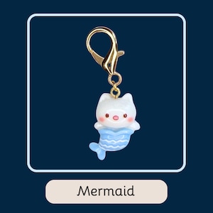 Puede incluir: Un clip de llavero dorado adjunto a un peque&ntilde;o dije de gato-sirena blanco con cola azul. El dije tiene nariz y mejillas rosas. La palabra "Mermaid" est&aacute; escrita en una etiqueta rectangular blanca debajo.
