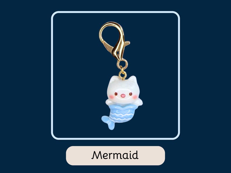 Puede incluir: Un clip de llavero dorado adjunto a un peque&ntilde;o dije de gato-sirena blanco con cola azul. El dije tiene nariz y mejillas rosas. La palabra "Mermaid" est&aacute; escrita en una etiqueta rectangular blanca debajo.
