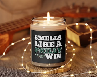 Può includere: Una candela marrone con un'etichetta nera che recita "Smells Like A Philly Win". L'etichetta ha un logo verde e bianco di un uccello con un pallone da calcio.