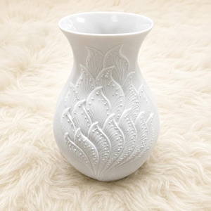 May include: A white ceramic vase with a flared rim and a rounded body. The front surface features a raised, embossed pattern of overlapping, stylized leaf or petal shapes, each detailed with fine lines and small, raised dots along the edges.