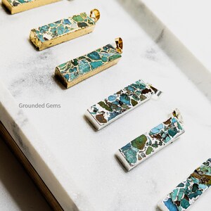 May include: Five turquoise and gold bar pendants with a gold chain. The pendants are made of a natural stone with a marbled pattern. The text "Grounded Gems" is visible in the image.