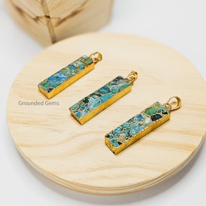 May include: Three gold-plated pendants with a rectangular shape and a blue and green marbled pattern. The pendants are displayed on a wooden surface.