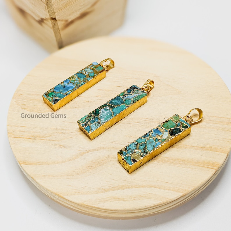 May include: Three gold-plated pendants with a rectangular shape and a blue and green marbled pattern. The pendants are displayed on a wooden surface.