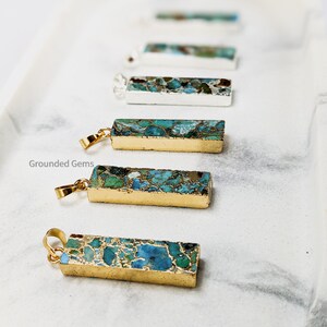 May include: A set of six gold-plated bar pendants with a blue and green marbled pattern. The pendants are arranged in a row on a white surface. The text "Grounded Gems" is visible in the lower left corner of the image.