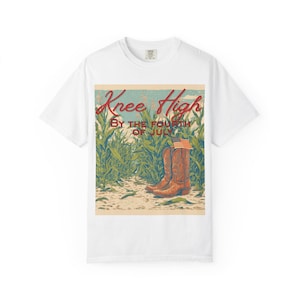 Op de afbeelding: Wit t-shirt met een vintage afbeelding. Het ontwerp toont bruine cowboylaarzen, een Amerikaanse vlag en de tekst "Knee High By The Fourth Of July" in rood en groen. De achtergrond toont een maïsveld.