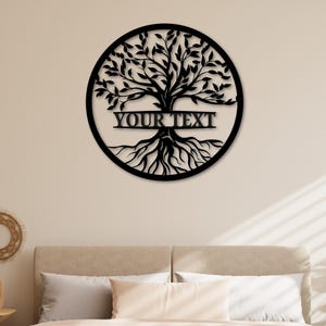 K&ouml;nnte beinhalten: Schwarze Metall-Wandkunst mit einem Lebensbaum und dem Text "YOUR TEXT" in einem horizontalen Banner. Der Baum befindet sich in einem kreisf&ouml;rmigen Rahmen. Die Wandkunst wird &uuml;ber einem Bett mit neutralfarbener Bettw&auml;sche gezeigt.