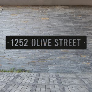 Könnte beinhalten: Schwarzes Metall-Adressschild mit dem Text "1252 OLIVE STREET" in einer Schablonenschrift. Das rechteckige Schild ist an einer grauen Steinmauer befestigt. Ein dekoratives Element für ein Haus oder Geschäft.