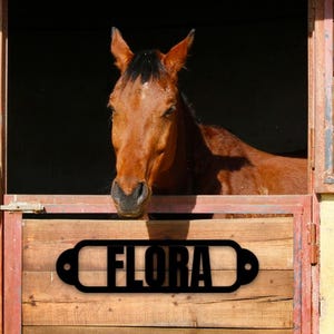 May include: A brown horse peeks out from a wooden stable. The stable door features a black metal sign that reads "FLORA". The roof is made of red tiles. The scene is bathed in sunlight, creating a warm, inviting atmosphere.