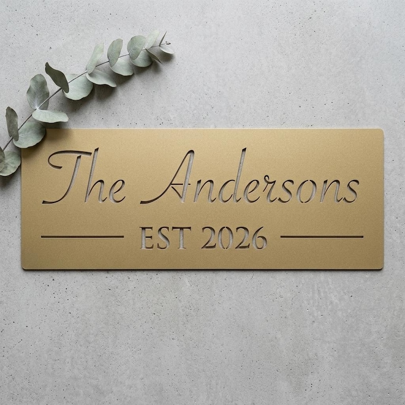 May include: A rectangular gold-coloured metal sign featuring the laser-cut text "The Andersons" in a cursive font above the text "EST 2026" flanked by two horizontal lines. A sprig of green eucalyptus leaves is positioned at the top left corner.