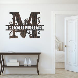 Peut inclure: Une enseigne murale en bois avec la lettre "M" et le nom "MCCULLOCH" d&eacute;coup&eacute;s. L'enseigne a une finition brun fonc&eacute; vein&eacute;e de bois et des ornements d&eacute;coratifs. Elle est fix&eacute;e sur un mur blanc.