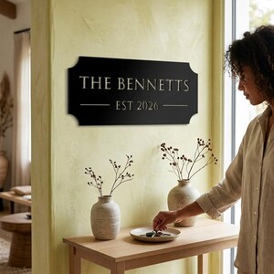 Pode incluir: Uma placa de parede retangular preta com cantos arredondados apresentando o texto na cor dourada THE BENNETTS e EST 2026. A placa est&aacute; montada numa parede amarelo-claro sobre uma mesa de consola de madeira com dois vasos de cer&acirc;mica.