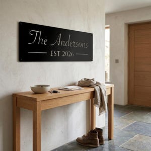 May include: A rectangular black metal wall sign featuring the white text The Andersons and EST 2026 with horizontal lines on either side of the date. The sign is mounted on a wall and secured with four visible screws at the corners of the panel.