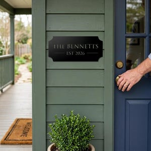 Pode incluir: Uma placa retangular de metal preto com bordas decorativas recortadas montada em uma parede verde. A placa apresenta o texto THE BENNETTS em uma fonte serifada, com o texto EST 2026 centralizado abaixo, separado por duas linhas horizontais.