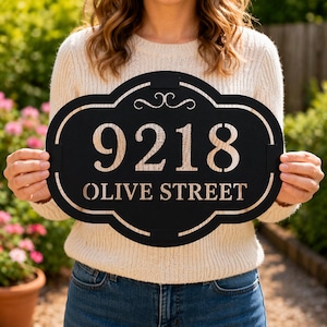 May include: Black metal address sign displaying the numbers "9218" and the words "OLIVE STREET". The sign has a decorative border. The sign is held by a person.