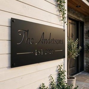 May include: A rectangular matte black metal sign mounted on a white horizontal siding wall. The sign features laser-cut lettering that reads The Andersons and EST 2026, with a horizontal line separating the text. It is secured by four circular fasteners.