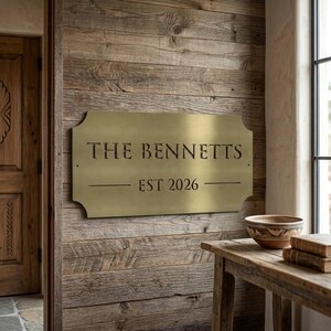 Pode incluir: Uma placa retangular de metal na cor lat&atilde;o com cantos arredondados e recortes decorativos. A placa apresenta o texto gravado THE BENNETTS e EST 2026, separados por linhas horizontais. Est&aacute; montada numa parede de t&aacute;buas de madeira.