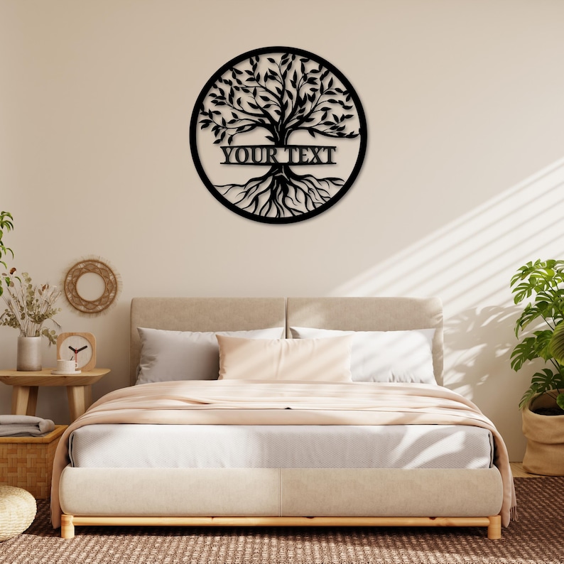 K&ouml;nnte beinhalten: Schwarze Metall-Wandkunst mit einem Lebensbaum und dem Text "YOUR TEXT" in einem horizontalen Banner. Der Baum befindet sich in einem kreisf&ouml;rmigen Rahmen. Die Wandkunst wird &uuml;ber einem Bett mit neutralfarbener Bettw&auml;sche gezeigt.