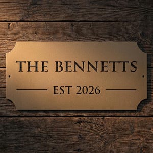Pode incluir: Uma placa retangular de metal na cor bronze com cantos arredondados e dois pequenos orif&iacute;cios de montagem nas laterais. A placa apresenta o texto cortado a laser THE BENNETTS em fonte serifada, com o texto EST 2026 centralizado abaixo entre duas linhas.