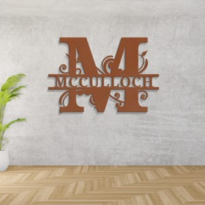 Peut inclure: Une enseigne murale en m&eacute;tal de couleur rouille avec la lettre "M" et le nom "MCCULLOCH" en dessous. Le design comprend des ornements d&eacute;coratifs. L'enseigne est fix&eacute;e sur un mur gris au-dessus d'un sol en bois.