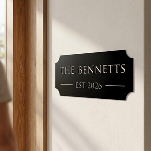 Pode incluir: Uma placa de parede retangular preta com cantos decorativos recortados, apresentando o texto prateado THE BENNETTS e EST 2026 centralizado na superf&iacute;cie, com linhas horizontais ladeando o ano para um visual limpo e equilibrado.