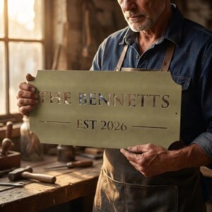 Pode incluir: Uma placa retangular de metal com cantos arredondados apresentando o texto recortado THE BENNETTS e EST 2026. A placa tem um acabamento verde-oliva fosco e &eacute; segurada horizontalmente. As letras s&atilde;o claras e centralizadas na pe&ccedil;a met&aacute;lica.