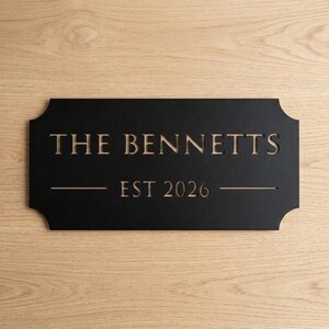 Pode incluir: Uma placa retangular preta com cantos arredondados apresentando o texto cortado a laser THE BENNETTS e EST 2026. As letras s&atilde;o recortadas para revelar a superf&iacute;cie de madeira clara por baixo, criando um visual de alto contraste.