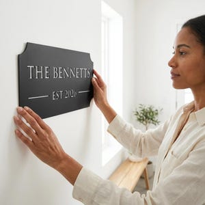 Pode incluir: Uma placa de parede retangular de metal preto com bordas curvas decorativas. A placa apresenta o texto cortado a laser THE BENNETTS em uma fonte serifada, seguido pelo texto EST 2026 centralizado entre duas linhas horizontais.