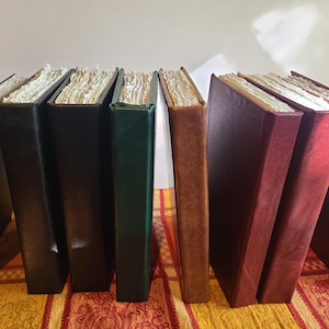 May include: A row of eight hardcover books standing upright, featuring textured covers in shades of black, dark green, brown, and deep red. Each book has visible, thick, off-white paper pages with deckled edges, creating a distinct layered appearance.