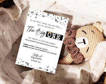 May include: A white invitation card with black text that reads "The Big One" and event details. The card is decorated with small black shapes. A beige crocheted teddy bear and a brown crocheted flower are next to the card.