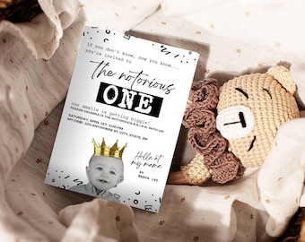 May include: A baby shower invitation with the text "The notorious ONE" and a baby wearing a gold crown. The invitation is on a white background with black accents and is accompanied by a beige crocheted teddy bear.
