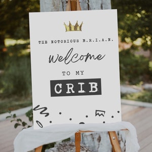K&ouml;nnte beinhalten: Ein wei&szlig;es Schild mit schwarzem Text, der "The Notorious B.R.I.A.N. welcome to my CRIB" lautet, mit einer goldenen Krone &uuml;ber dem Text. Das Schild steht auf einem Staffelei und hat schwarze Kritzel-Akzente am unteren Rand.