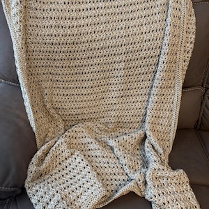May include: A handmade, light brown crochet blanket with a delicate open weave pattern. The blanket is draped over a dark brown sofa, showcasing its texture and design. The blanket's edges have a decorative border.