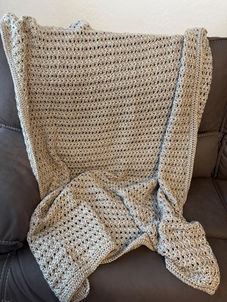 May include: A handmade, light brown crochet blanket with a delicate open weave pattern. The blanket is draped over a dark brown sofa, showcasing its texture and design. The blanket's edges have a decorative border.
