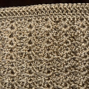 May include: Close-up of a hand-crocheted item in shades of tan and brown. The textured surface features a repeating pattern of raised stitches, creating a dimensional effect. The top edge has a decorative border.