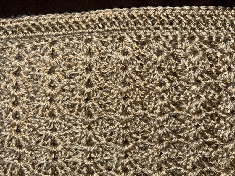 May include: Close-up of a hand-crocheted item in shades of tan and brown. The textured surface features a repeating pattern of raised stitches, creating a dimensional effect. The top edge has a decorative border.