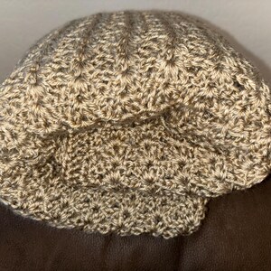 May include: A crocheted hat in shades of tan and brown. The hat has a textured, ribbed design on the crown and a folded brim. The handmade hat is likely made of a soft yarn, perfect for cooler weather.