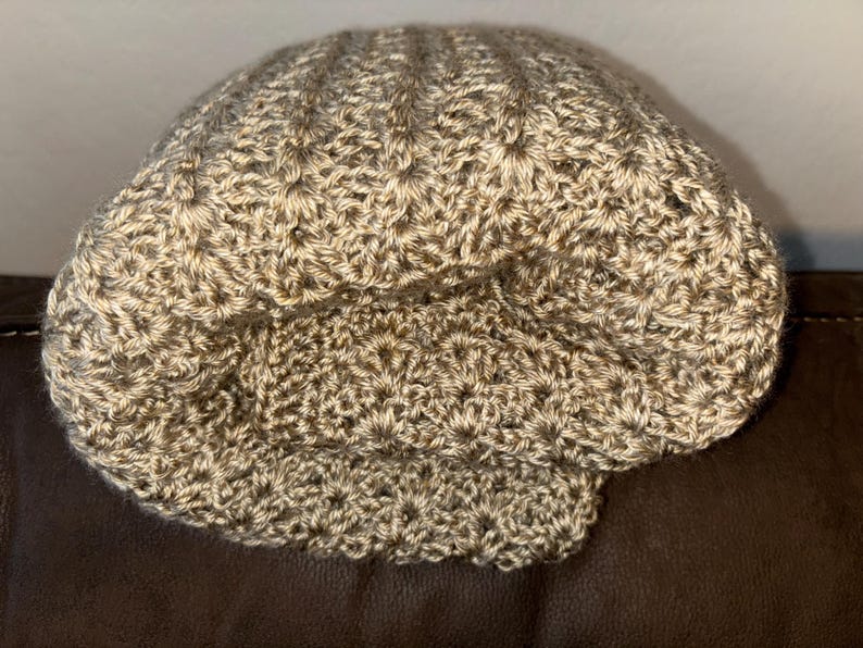 May include: A crocheted hat in shades of tan and brown. The hat has a textured, ribbed design on the crown and a folded brim. The handmade hat is likely made of a soft yarn, perfect for cooler weather.