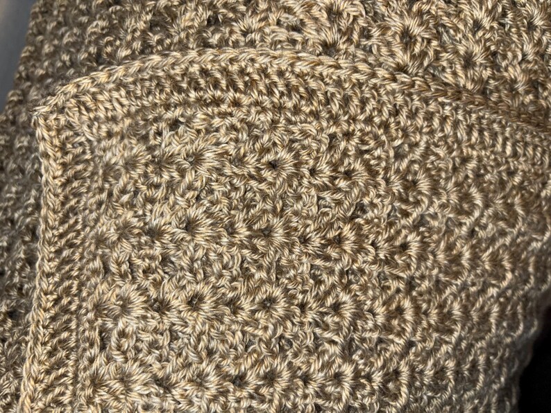 May include: Close-up of a hand-crocheted item in shades of tan and beige. The textured surface reveals a detailed stitch pattern, with a visible border. The item appears to be a blanket or shawl, showcasing the craft's intricate design.