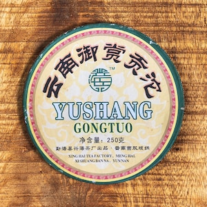 以下が含まれることがあります： 中国語の文字と「YUSHANG GONGTUO」の文字が緑と黒で書かれた丸いお茶のケーキ。お茶のケーキは緑色の縁とクリーム色の中心を持っています。「净含量: 250克」の文字も表示されています。