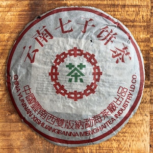 以下が含まれることがあります： 赤と緑のアクセントが付いた、丸い銀色の茶葉ケーキ。茶葉ケーキには、中国語の文字と中央に円形のデザインがあります。テキストには「China Yunnan Xishuangbanna Menghai Tea Industry LD. Co.」が含まれています。