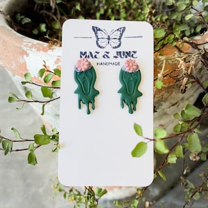 Könnte beinhalten: Ein Paar grüne Geisterohrringe mit rosa und lila Blumen oben. Die Ohrringe sind handgefertigt und befinden sich auf einer weißen Karte mit dem Text "Mae & June Handmade".