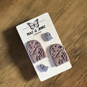 May include: A set of three purple polymer clay earrings. Two earrings feature a desert landscape with a cactus, mountains, and a crescent moon. The third earring is a ghost.