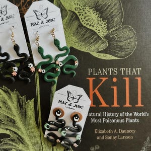 May include: Handmade snake earrings in black and green with floral accents. The earrings are displayed on white tags with the text "MAZ & JUNZ HANDMADE". The background includes a book cover with the title "PLANTS THAT KILL".
