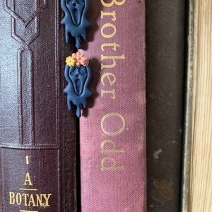 Könnte beinhalten: Zwei dunkelblaue, geisterförmige Ohrringe mit einem schreienden Gesicht und einer Blumenkrone aus rosa, gelben und orangefarbenen Blumen. Die Ohrringe befinden sich auf einem Regal mit Büchern dahinter.