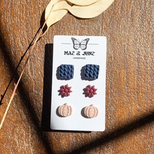 May include: A set of six polymer clay earrings on a white card with the text "Mae & June Handmade". The earrings are in three different designs: a blue knitted square, a burgundy flower, and a peach pumpkin with white polka dots.