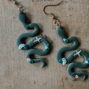May include: A pair of dark green snake-shaped earrings with white and blue floral accents. The earrings are made of polymer clay and have gold hooks.