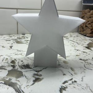 May include: A white, five-pointed star decoration. The star is elevated on a rectangular base. The star is made of a textured material and is set against a white tile background. The star is a decorative item.