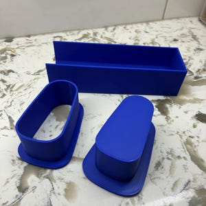 May include: A set of three blue plastic tools consisting of one long rectangular tray with an open top and two oval-shaped cutters or molds. The objects feature smooth, solid blue surfaces and are arranged on a light-colored surface.
