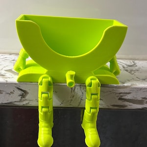May include: A bright lime green plastic holder featuring a semi-circular basin and two articulated legs that hang downward. The structure includes a small central protrusion and two arms positioned at the sides of the basin, creating a seated figure.