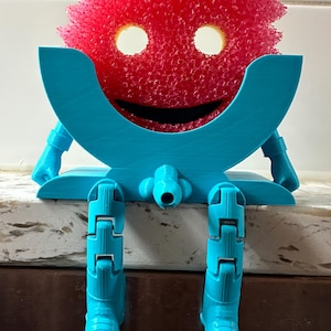 May include: A whimsical, 3D-printed blue sponge holder shaped like a smiling face with articulated legs. The holder cradles a bright pink, textured cleaning sponge, creating a playful and functional kitchen accessory.